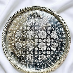 May include: A round, silver-colored metal tray with an intricate floral and geometric pattern. The tray's edge has a rope-like design. The detailed design is in a contrasting white color, creating a striking visual effect. The tray is likely used for serving or display.