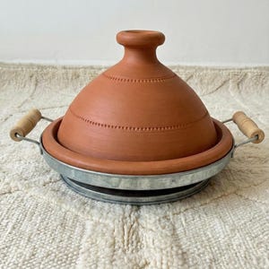 May include: A terracotta tagine pot with a conical lid and a metal base. The lid has a series of small, evenly spaced indentations. The pot has two wooden handles. The tagine is designed for cooking and serving Moroccan dishes.