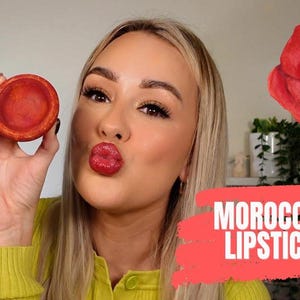 May include: A woman holds a small, round, terracotta-colored container of Moroccan lipstick. The text "MOROCCAN LIPSTICK" is displayed on a red background. A red poppy flower is in the background.