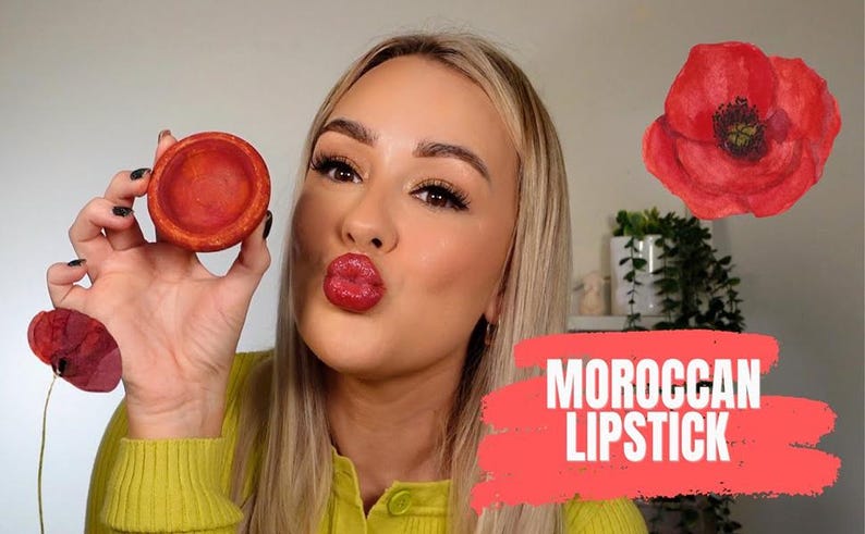 May include: A woman holds a small, round, terracotta-colored container of Moroccan lipstick. The text "MOROCCAN LIPSTICK" is displayed on a red background. A red poppy flower is in the background.