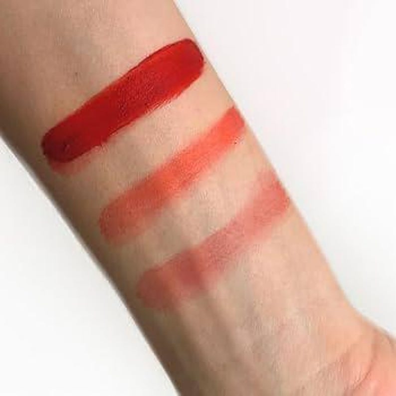 May include: A close-up of an arm with four swatches of red and orange lipstick. The top swatch is a vibrant, opaque red. The other three swatches are shades of orange and coral, with the bottom swatch being the lightest.