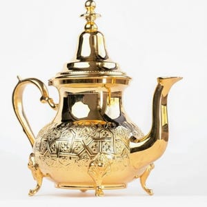 Op de afbeelding: Een goudkleurige theepot met ingewikkelde reliëfpatronen. De theepot heeft een gebogen tuit, een handvat en een deksel met een decoratieve knop. Hij staat op drie kleine voetjes en het ontwerp is sierlijk en elegant.