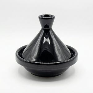 Op de afbeelding: Een zwarte keramische tajine-pot met een conisch deksel en een brede, ondiepe basis. De tajine heeft een glanzende afwerking en reflecteert licht. Traditioneel ontwerp voor Marokkaans koken.