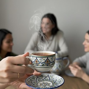 Puede incluir: Una taza de cerámica blanca con un asa negra y el texto "But First, Coffee" en negrita negra. La taza está llena de un líquido oscuro, probablemente café, y se encuentra sobre una superficie clara.