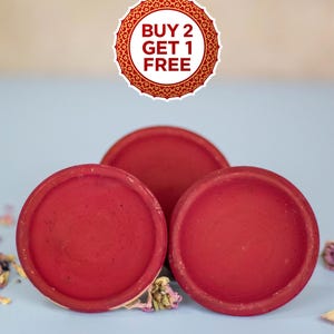 May include: Three round, red terracotta dishes are arranged on a light blue surface. The dishes have a slightly rough texture and a deep red color. A circular sticker with the text "BUY 2 GET 1 FREE" is at the top.