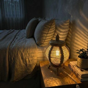 Könnte beinhalten: Dekorative Nachttischlampe mit geflochtenem Seil-Schirm und Holzsockel. Die Lampe ist eingeschaltet und wirft ein warmes Licht auf einen Nachttisch aus Holz. Eine kleine Topfpflanze und Bücher befinden sich ebenfalls auf dem Nachttisch.