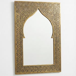 Pode incluir: Espelho retangular dourado com topo arqueado. A moldura apresenta padrões intrincados em relevo. O espelho é montado numa parede branca, exibindo um design decorativo e ornamentado, adequado para decoração de interiores.