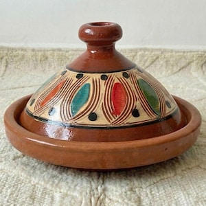 Op de afbeelding: Een bruine keramische tajinepot met een deksel. Het deksel is versierd met kleurrijke patronen van rood, groen en blauw. De pot is rond met een brede basis en een conisch deksel, ideaal om in te koken en te serveren. De tajine is een traditioneel Noord-Afrikaans kookvat.