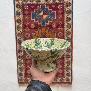 Puede incluir: Un cuenco con pedestal crema, salpicado de pintura verde y con borde festoneado. El cuenco se sostiene frente a una alfombra estampada roja y beige.