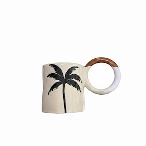 Könnte beinhalten: Cremefarbene Keramiktasse mit einem schwarzen Palmenmuster. Der runde Griff ist oben braun und unten weiß. Eine einzigartige Tasse für Tee oder Kaffee, die jeder Küche einen Hauch von Stil verleiht.