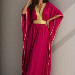 Può includere: Un abito caftano color magenta con finiture dorate e una cintura ricamata in oro. L'abito ha uno scollo a V e maniche ampie. La modella indossa l'abito davanti a un muro grigio.