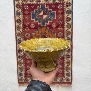 Puede incluir: Un cuenco de cerámica amarillo-verdoso hecho a mano con un borde festoneado, sostenido frente a una alfombra estampada. El cuenco tiene una base de pedestal y una superficie texturizada, mostrando una estética rústica. La alfombra presenta un fondo rojo con diseños geométricos.