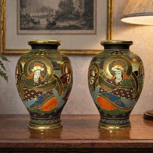 Könnte beinhalten: Zwei verzierte, antike japanische Vasen mit goldenen Akzenten und detaillierten Kunstwerken, die Figuren in traditioneller Kleidung darstellen. Die Vasen haben einen schwarzen Rand und Sockel und stehen auf einer Holzoberfläche.