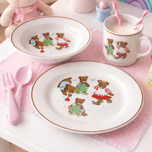 Könnte beinhalten: Ein Kindergeschirr-Set mit Teller, Schüssel und Becher, jeweils mit einer Illustration eines braunen Bären. Das Set enthält rosa Besteck und liegt auf einer rosa gepunkteten Tischdecke. Ein Stoffhase und Blumen sind im Hintergrund.