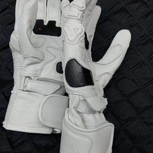 May include: A pair of white motorcycle gloves with black and red accents. The gloves have reinforced knuckles and wrist straps for added protection. The design includes padded sections and a textured palm for grip. The gloves are made of leather and have a sporty aesthetic.