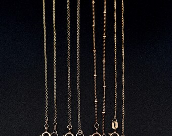 May include: Seven gold chains of varying lengths and styles hanging against a black background. The chains are all made of a delicate, fine gauge metal.