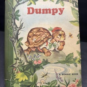 Puede incluir: Libro infantil vintage titulado "Dumpy". La portada presenta una ilustración de una tortuga con sombrero y camisa, sentada en una roca. El fondo incluye un estanque, flores y un árbol. El texto "A Bonnie Book" está en la parte inferior.