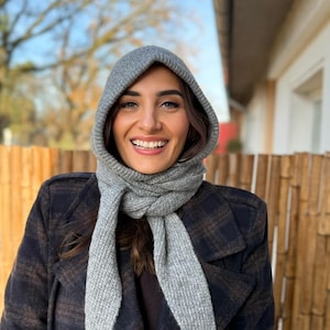 Może przedstawiać: Szary zestaw z dzianiny z kapturem i szalikiem. Kaptur jest podniesiony, oprawiając twarz, a szalik jest owinięty wokół szyi. Osoba ma na sobie ciemny płaszcz w kratę. Tło przedstawia rozmyty widok na zewnątrz.