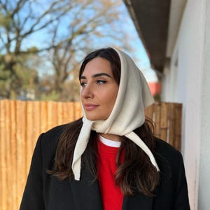 Può includere: Una donna indossa un foulard color crema con lunghe cravatte, un maglione rosso e un cappotto nero. Il foulard è legato sotto il mento. Lo sfondo comprende una staccionata di legno e un albero.