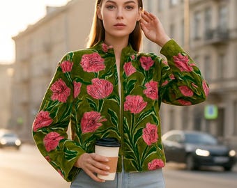 May include: A green velvet jacket with a pink floral pattern. The jacket has a stand-up collar and a zipper closure. The model is wearing light blue jeans and holding a coffee cup. The background is a city street.