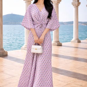 May include: A light purple maxi dress with a floral pattern, a V-neck, and a tie waist. The dress has short, wide sleeves and is paired with a straw hat and a small white handbag. The background features a seaside view.