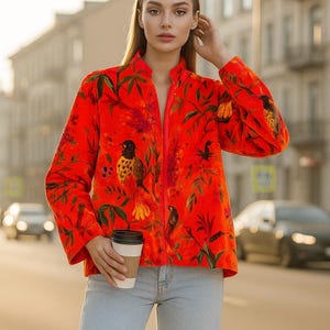 Può includere: Giacca rosso vivo con stampa botanica e uccelli. La giacca ha un collo alto e una cerniera a tutta lunghezza. Il design include foglie verdi e uccelli colorati. La persona tiene in mano una tazza di caffè e indossa jeans azzurri.
