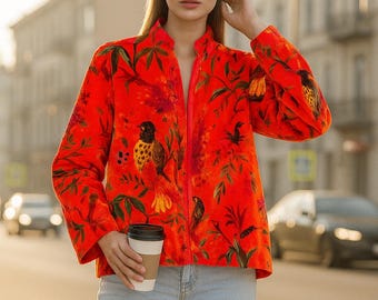 May include: A vibrant red jacket with a botanical and bird print. The jacket features a high collar and a full-length zipper. The design includes green leaves and colorful birds. The person is holding a coffee cup and wearing light blue jeans.