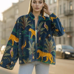 May include: A dark blue jacket with a jungle-themed pattern featuring yellow leopards, toucans, and palm trees. The jacket has a mandarin collar and a front zip. The model is wearing light blue jeans.