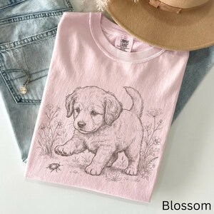May include: A pale pink t-shirt with a detailed sketch of a puppy interacting with a bug. The puppy is surrounded by floral elements. The text "Blossom" is at the bottom right corner of the shirt.