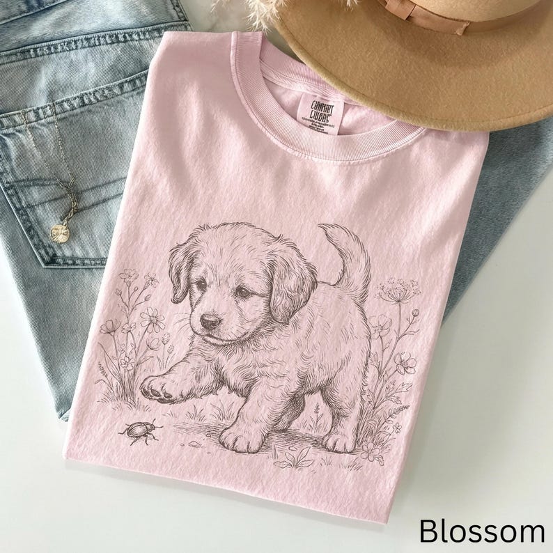 May include: A pale pink t-shirt with a detailed sketch of a puppy interacting with a bug. The puppy is surrounded by floral elements. The text "Blossom" is at the bottom right corner of the shirt.