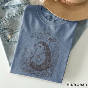 Puede incluir: Camiseta azul vaquero con una ilustraci&oacute;n marr&oacute;n de un erizo sosteniendo un paraguas de hojas. El erizo est&aacute; rodeado de plantas y flores. Un sombrero color canela y unos vaqueros azul claro est&aacute;n en el fondo.