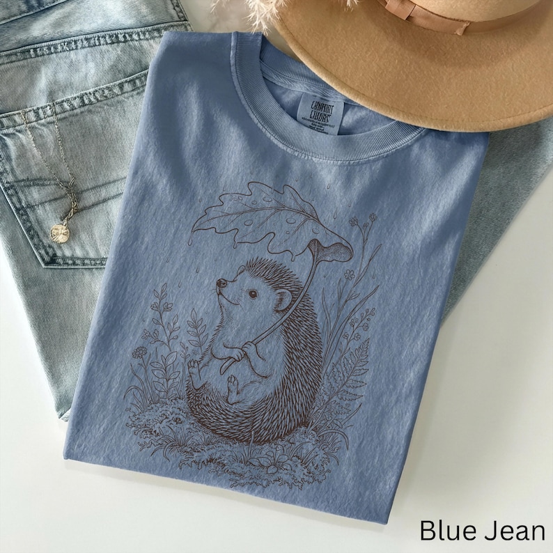 Puede incluir: Camiseta azul vaquero con una ilustraci&oacute;n marr&oacute;n de un erizo sosteniendo un paraguas de hojas. El erizo est&aacute; rodeado de plantas y flores. Un sombrero color canela y unos vaqueros azul claro est&aacute;n en el fondo.