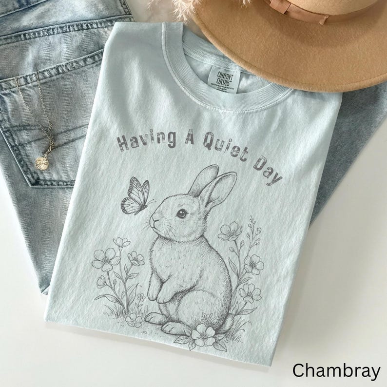 May include: A light blue t-shirt with the text "Having A Quiet Day" above a detailed illustration of a rabbit and butterfly. The shirt is made of soft material and is paired with light wash denim jeans and a tan hat.