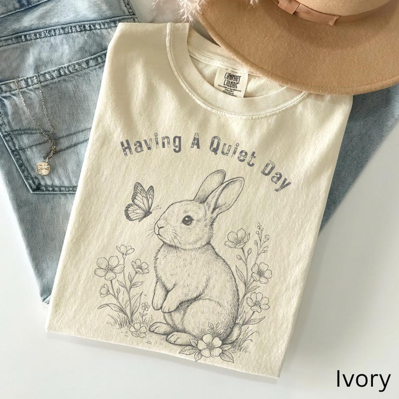 May include: Ivory-colored t-shirt with the text "Having A Quiet Day" above a detailed illustration of a rabbit and butterfly. The rabbit is surrounded by flowers. The shirt is made of a soft material.