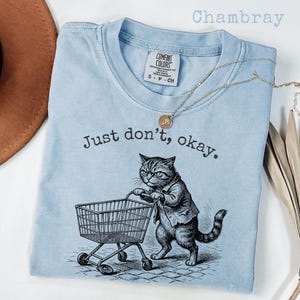 May include: Light blue t-shirt with the text "Just don't, okay." and a graphic of a cat pushing a shopping cart. The shirt has a crew neck and short sleeves. A gold necklace is draped across the shirt.