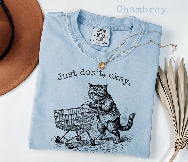 May include: Light blue t-shirt with the text "Just don't, okay." and a graphic of a cat pushing a shopping cart. The shirt has a crew neck and short sleeves. A gold necklace is draped across the shirt.