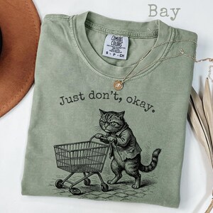 May include: A sage green t-shirt with the text "Just don't, okay." and a graphic of a cat pushing a shopping cart. The cat is wearing a jacket. A gold necklace is draped across the shirt. The shirt has a Comfort Colors tag.