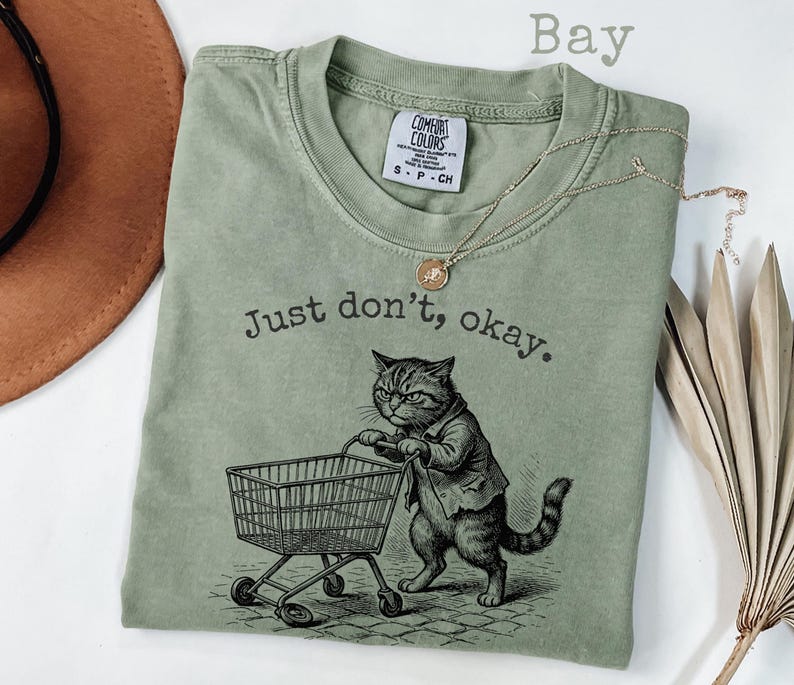May include: A sage green t-shirt with the text "Just don't, okay." and a graphic of a cat pushing a shopping cart. The cat is wearing a jacket. A gold necklace is draped across the shirt. The shirt has a Comfort Colors tag.