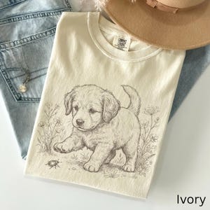 May include: Ivory-coloured t-shirt with a detailed illustration of a puppy and a beetle amongst floral elements. The shirt has a classic crew neck and is made of a soft, comfortable material. The word "Ivory" is printed at the bottom right.