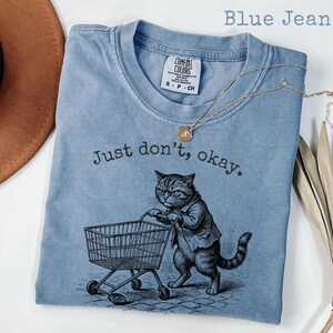 May include: Blue jean colored t-shirt with the text "Just don't, okay." and a graphic of a cat pushing a shopping cart. The shirt has a crew neck and short sleeves. A gold necklace with a pendant is draped across the shirt.