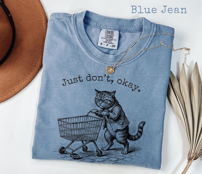 May include: Blue jean colored t-shirt with the text "Just don't, okay." and a graphic of a cat pushing a shopping cart. The shirt has a crew neck and short sleeves. A gold necklace with a pendant is draped across the shirt.