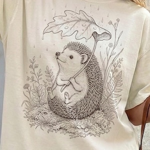 Puede incluir: Camiseta color crema con una ilustraci&oacute;n caprichosa de un erizo sentado en la hierba, sosteniendo un paraguas de hojas. El dibujo en blanco y negro incluye gotas de lluvia y plantas circundantes, creando una escena natural encantadora.