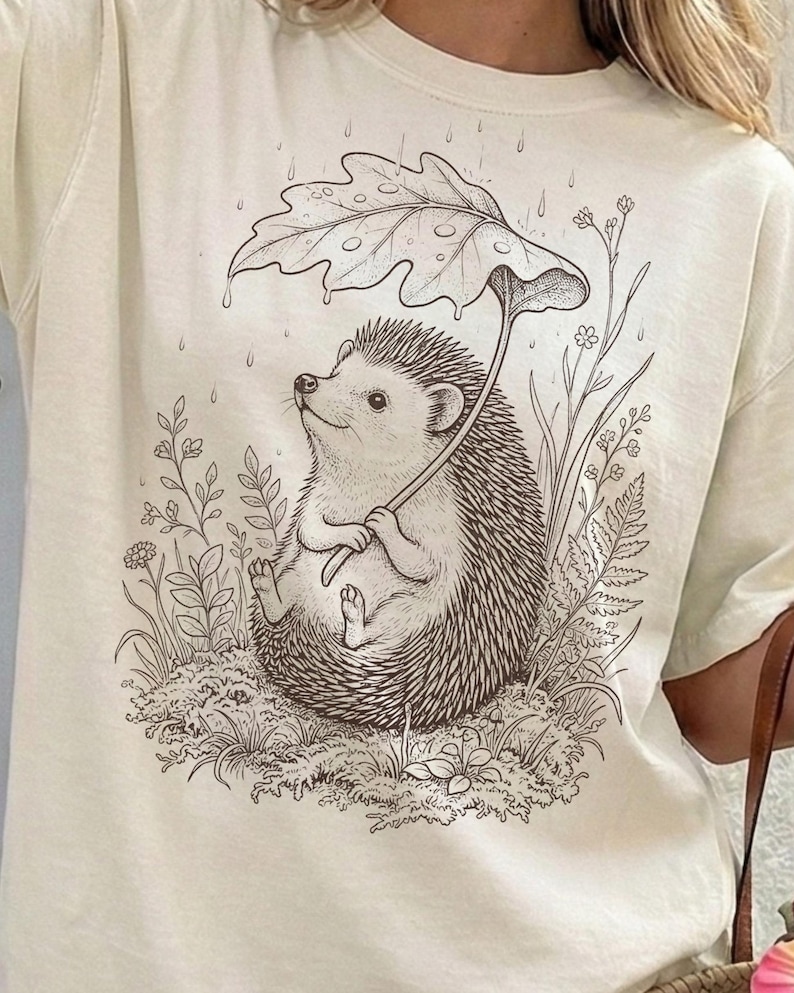 Puede incluir: Camiseta color crema con una ilustraci&oacute;n caprichosa de un erizo sentado en la hierba, sosteniendo un paraguas de hojas. El dibujo en blanco y negro incluye gotas de lluvia y plantas circundantes, creando una escena natural encantadora.