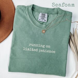May include: Seafoam green t-shirt with the phrase "running on limited patience" in white. A gold necklace with a pendant rests on the shirt. The shirt is folded, and a brown hat is partially visible.