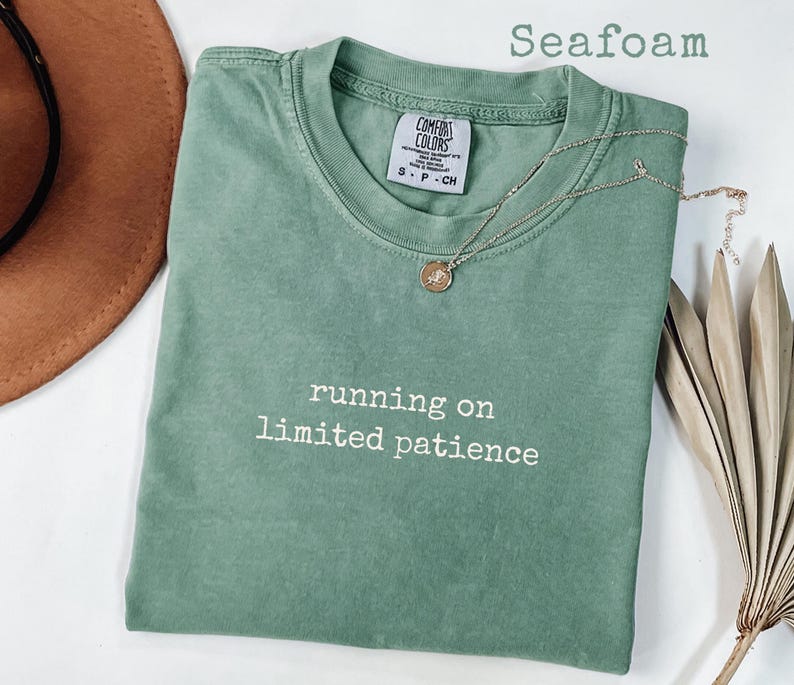 May include: Seafoam green t-shirt with the phrase "running on limited patience" in white. A gold necklace with a pendant rests on the shirt. The shirt is folded, and a brown hat is partially visible.