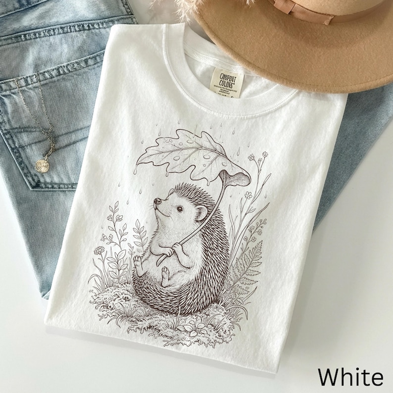 Puede incluir: Camiseta blanca con una ilustraci&oacute;n detallada de un erizo sosteniendo un paraguas de hojas, rodeado de plantas. La camiseta Comfort Colors tiene cuello redondo y mangas cortas. La palabra "White" est&aacute; impresa en la parte inferior derecha.