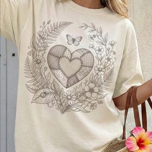 Peut inclure: T-shirt crème avec un motif de cœur détaillé composé de motifs patchwork. Le cœur est entouré d'une couronne de fleurs, de fougères et d'un papillon. Le motif est dans les tons de gris et de marron.