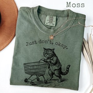 May include: Moss green t-shirt with a graphic of a cat pushing a shopping cart, and the text "Just don't, okay." A gold necklace is draped across the shirt. The shirt is made of a soft material.