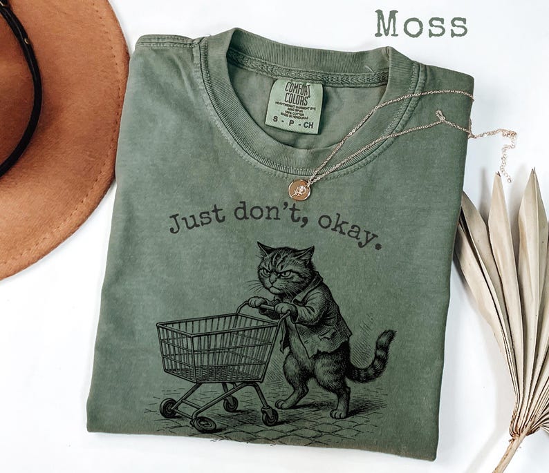 May include: Moss green t-shirt with a graphic of a cat pushing a shopping cart, and the text "Just don't, okay." A gold necklace is draped across the shirt. The shirt is made of a soft material.