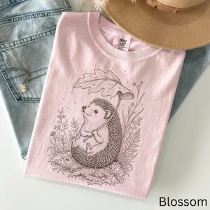 Puede incluir: Una sudadera Comfort Colors rosa claro con una ilustraci&oacute;n detallada de un erizo sosteniendo un paraguas de hojas. El erizo est&aacute; rodeado de plantas y flores. La palabra "Blossom" est&aacute; impresa en la esquina inferior derecha. Un sombrero color canela y unos vaqueros azules est&aacute;n en el fondo.
